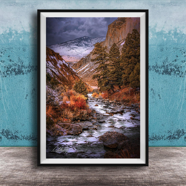 The image is a framed photograph of a scenic mountain landscape with a winding river flowing through it. The mountains are covered in snow, and the sky is filled with clouds, creating a dramatic and moody atmosphere. The photograph is framed and displayed on a wooden surface, with a blue wall in the background.