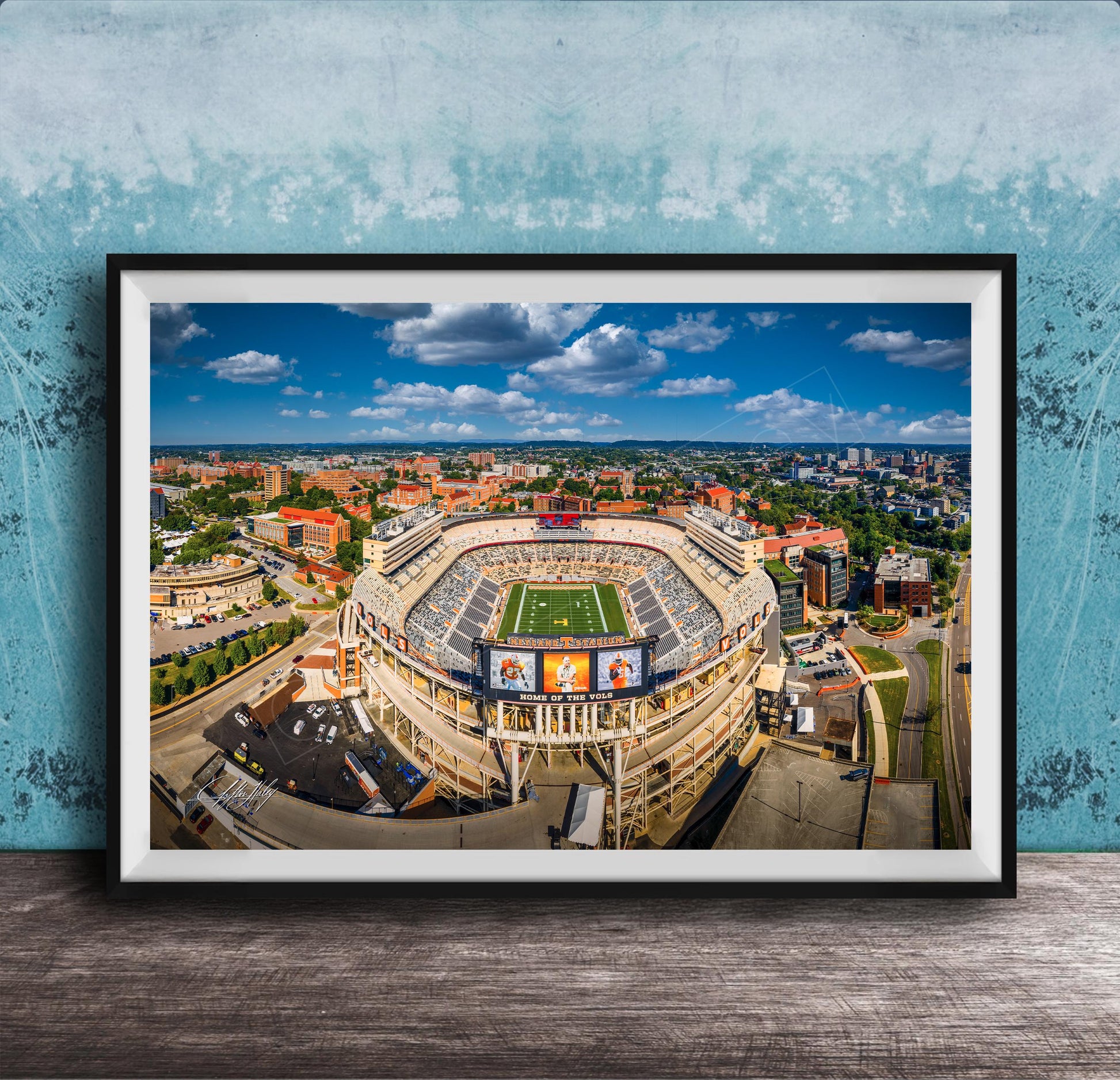An aerial view of a large sports stadium surrounded by a cityscape, with a clear blue sky and fluffy white clouds overhead.