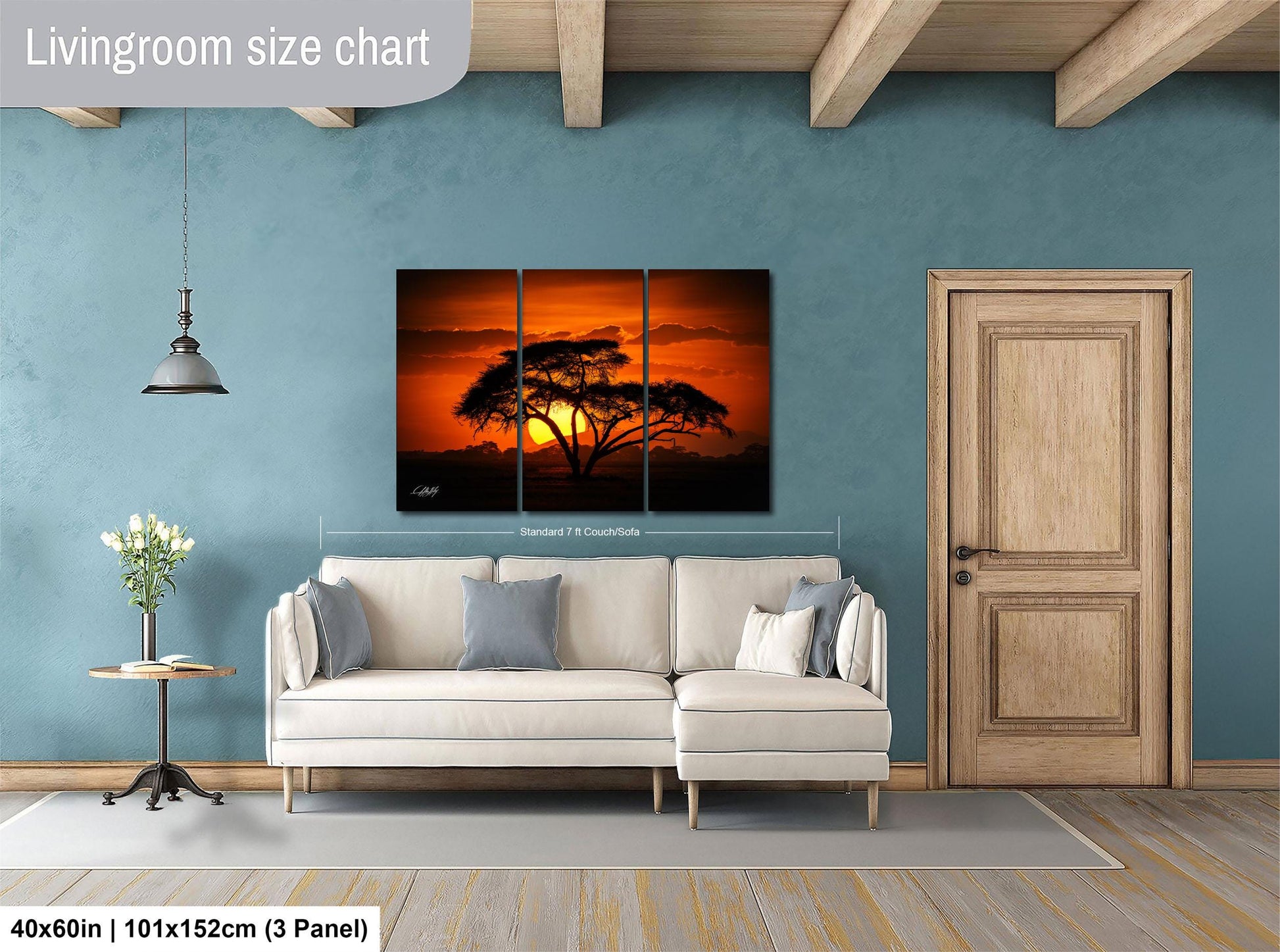 The image is a living room with a large, three-panel landscape painting depicting a sunset over a tree. The painting is hung on the wall above a white sofa, and there is a wooden door and a small table with a vase of flowers nearby. The room has a blue wall and wooden beams, creating a cozy and inviting atmosphere.