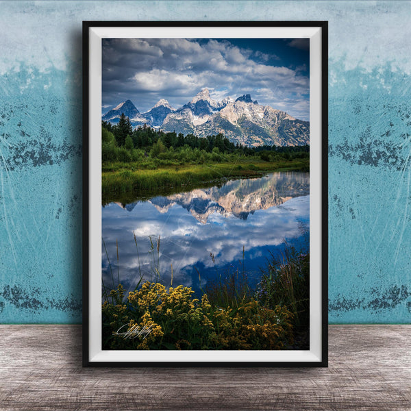 A framed photograph of a serene landscape with a mountain range reflected in a calm lake, surrounded by lush greenery and vibrant yellow flowers.