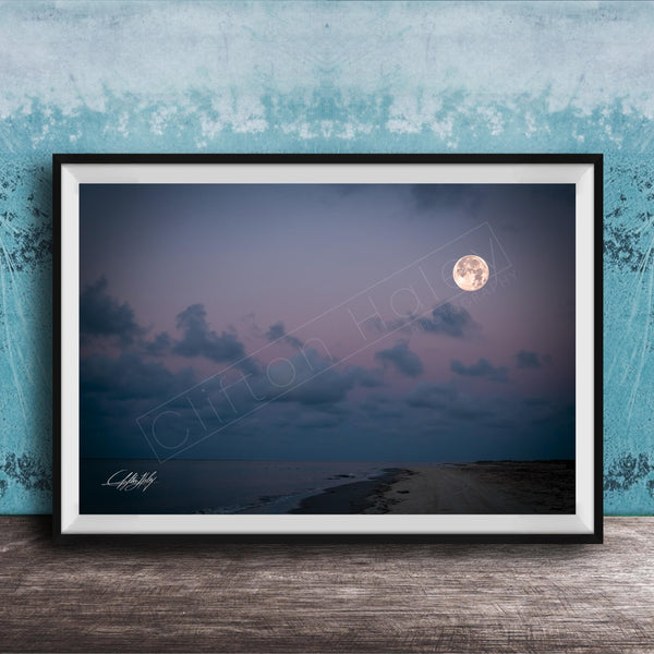 A framed photograph of a serene beach at night, with a full moon rising over the ocean and a cloudy sky.