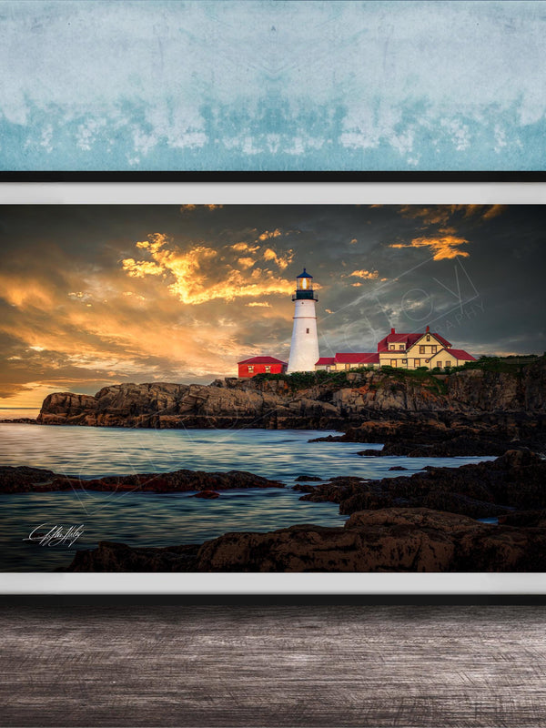 A framed photograph depicting a lighthouse on a rocky coastline at sunset, with the sky filled with clouds and the ocean in the background.
