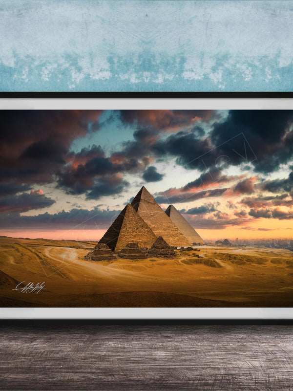 A framed photograph of the iconic pyramids of Giza against a dramatic sky at sunset, with the sun setting behind the sand dunes.