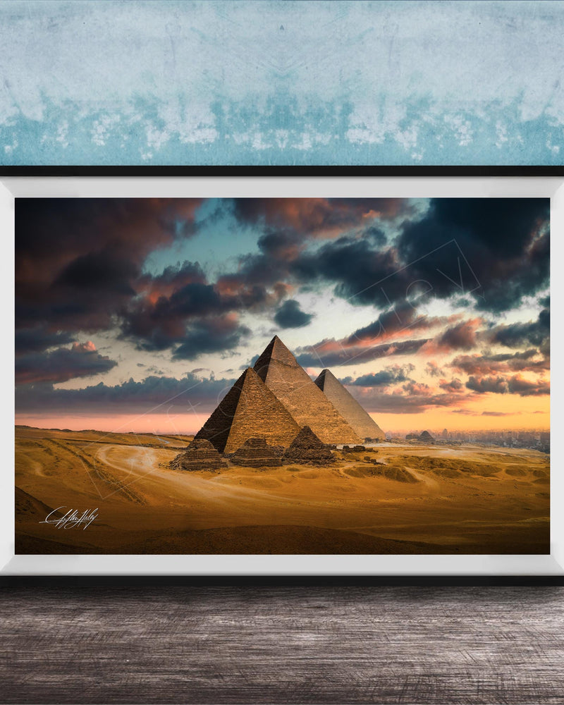 A framed photograph of the iconic pyramids of Giza against a dramatic sky at sunset, with the sun setting behind the sand dunes.