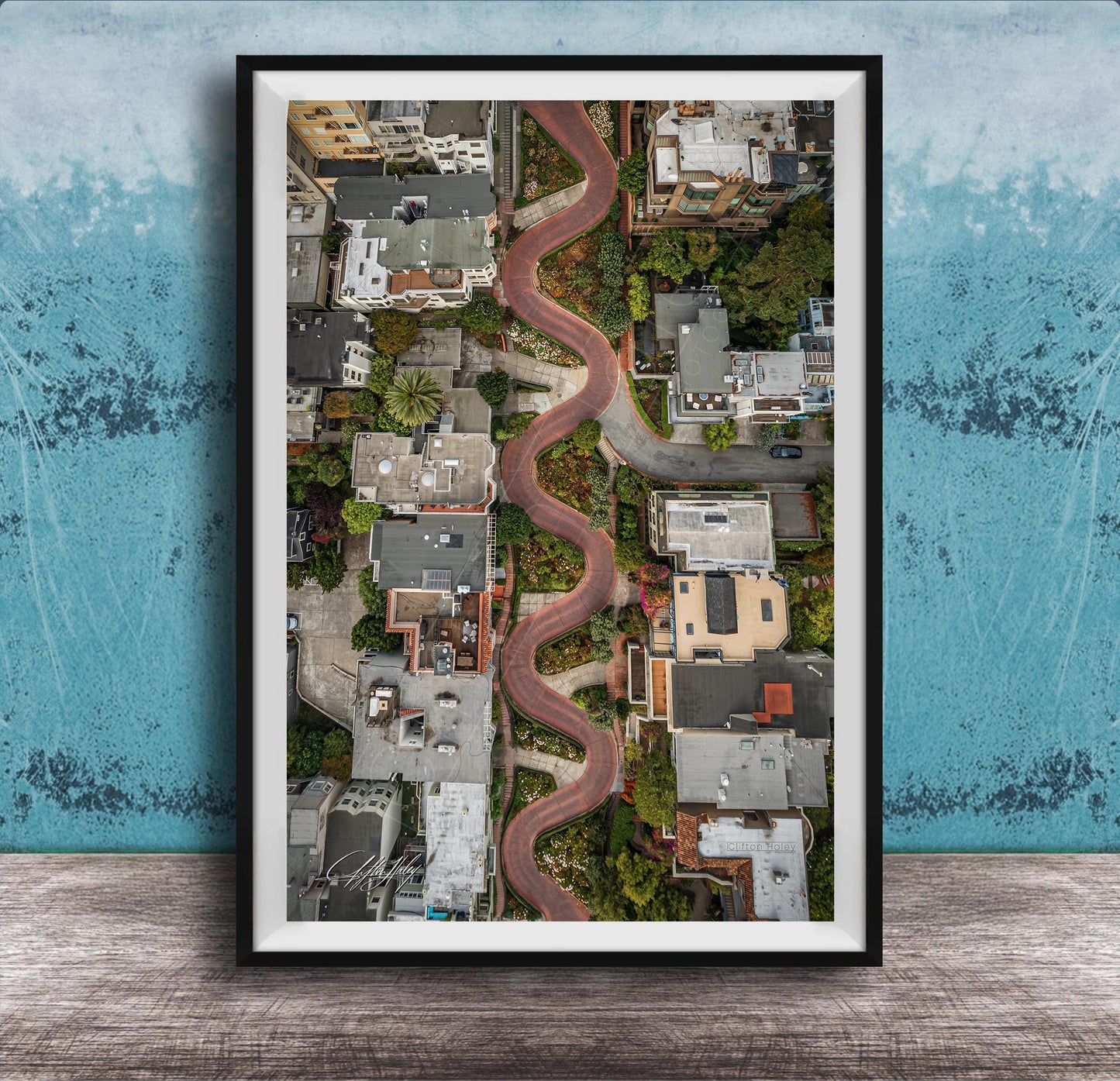 The image is an aerial view of a winding road that curves through a cityscape, with buildings and greenery lining the sides of the road.