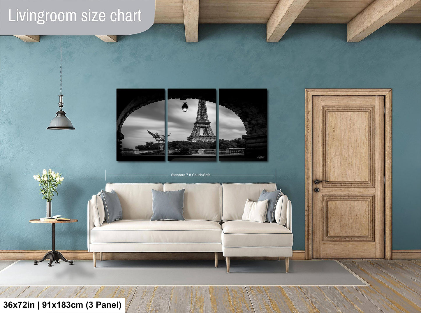 The image is a living room with a large wall art depicting the Eiffel Tower. The room has a white sofa, a wooden door, and a wooden table with a vase of flowers. The wall art is a collage of three panels, and the dimensions are provided as 36 inches by 21 inches by 3 panels.