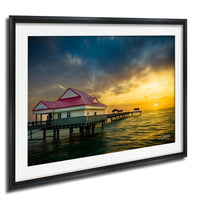 A framed photograph depicting a wooden pier with a red-roofed building, set against a vibrant sunset sky over the ocean.