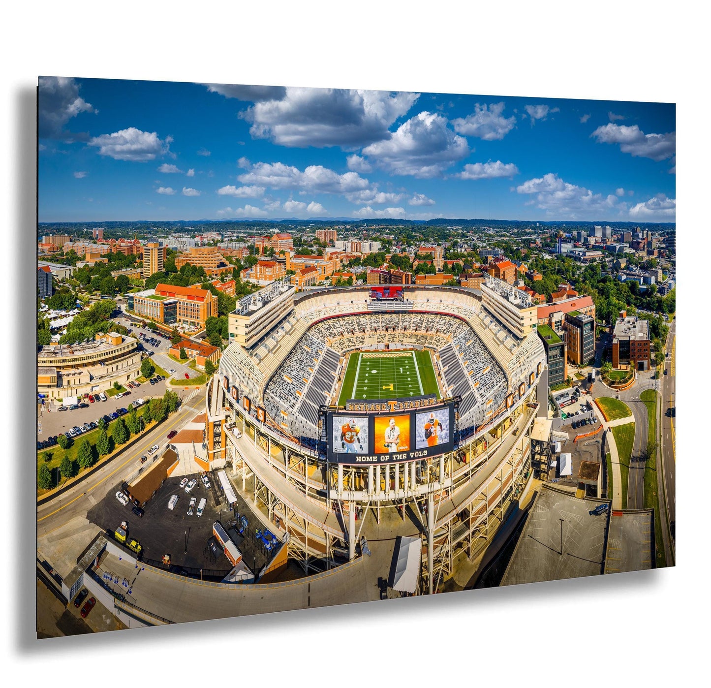 An aerial view of a large sports stadium surrounded by a cityscape, with a clear blue sky and scattered clouds above.