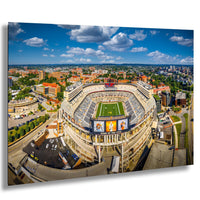An aerial view of a large sports stadium surrounded by a cityscape, with a clear blue sky and scattered clouds above.