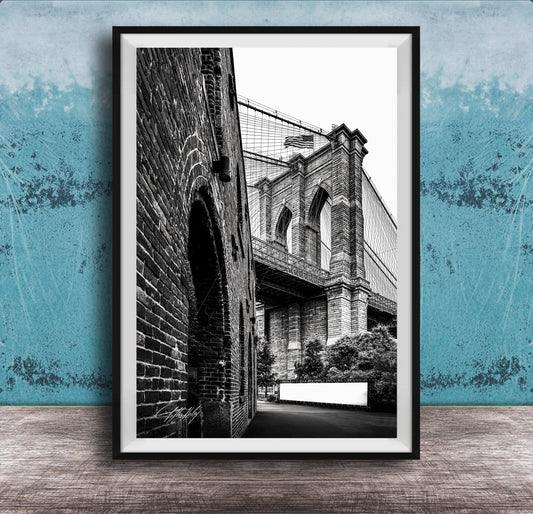 A black and white photograph of the Brooklyn Bridge, with a brick archway and a white sign visible in the foreground.