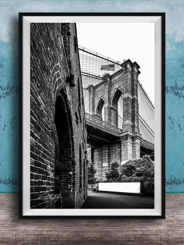 A black and white photograph of the Brooklyn Bridge, with a brick archway and a white sign visible in the foreground.