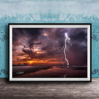 A framed photograph depicting a dramatic lightning storm over a beach at sunset, with the sky filled with vibrant colors and the lightning bolt striking the horizon.