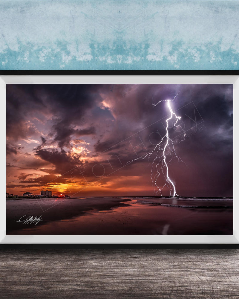 A framed photograph depicting a dramatic lightning storm over a beach at sunset, with the sky filled with vibrant colors and the lightning bolt striking the horizon.