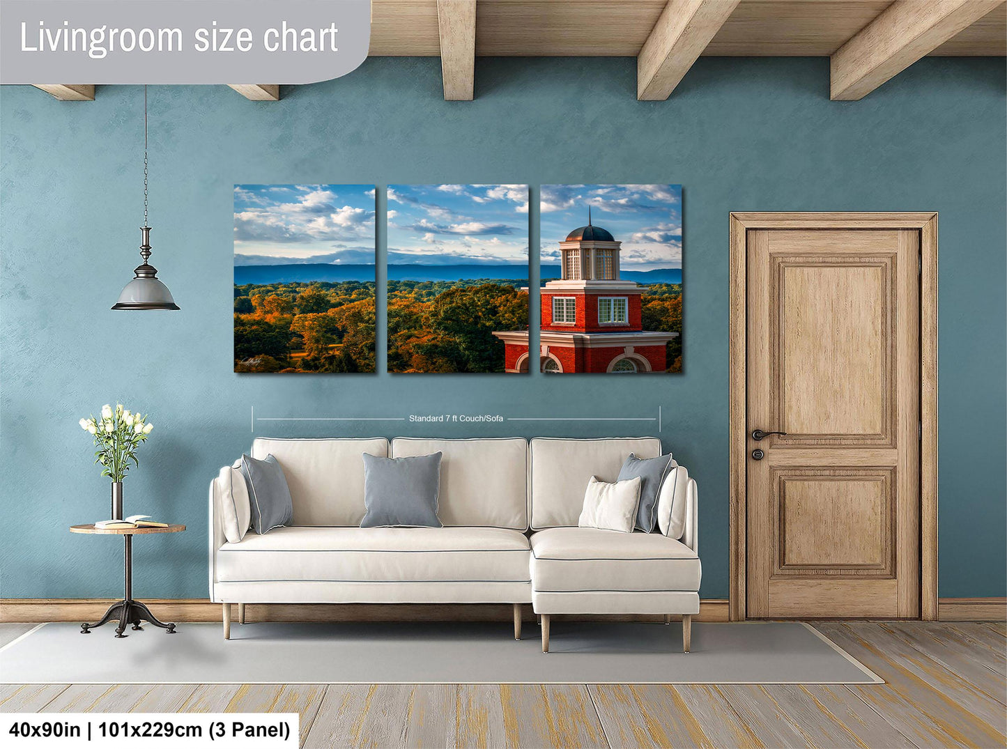 The image is a living room with a large, three-panel wall art depicting a scenic landscape with a red building in the background. The wall art is mounted above a white sofa, and there is a wooden door and a small table with a vase of flowers nearby. The living room appears to be well-lit and spacious, with a wooden floor and a ceiling with exposed beams.