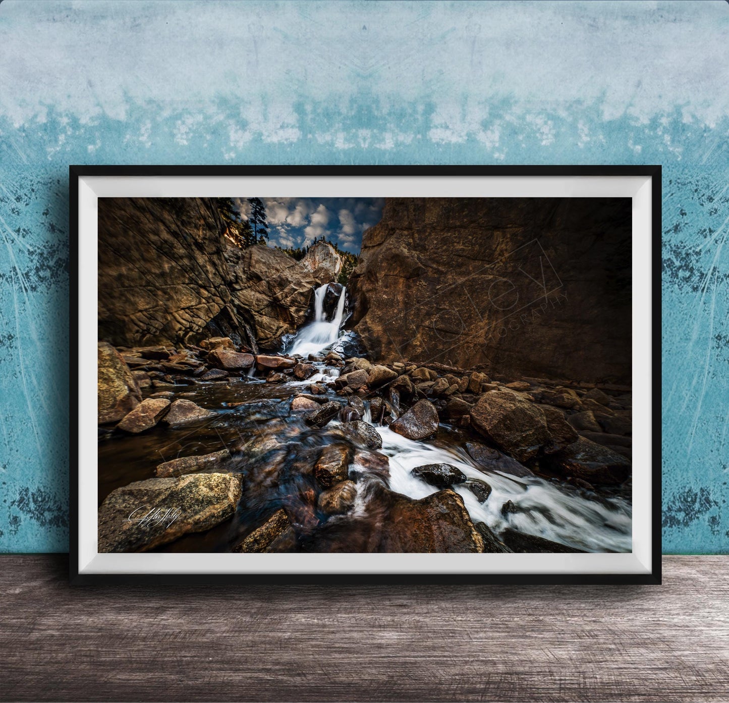 A framed photograph of a rocky waterfall cascading over rocks, set against a blue wall with a textured pattern.