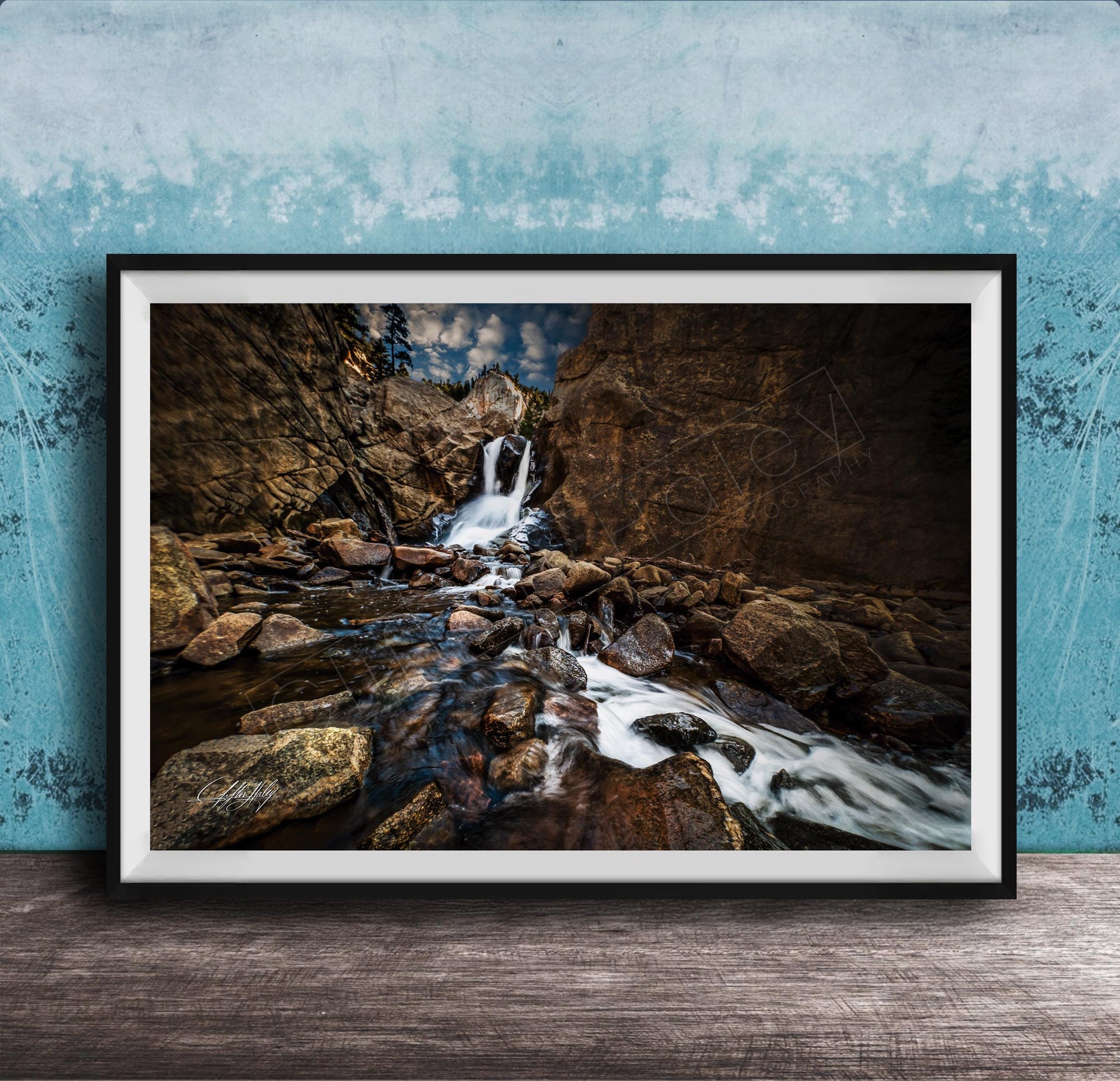 A framed photograph of a rocky waterfall cascading over rocks, set against a blue wall with a textured pattern.