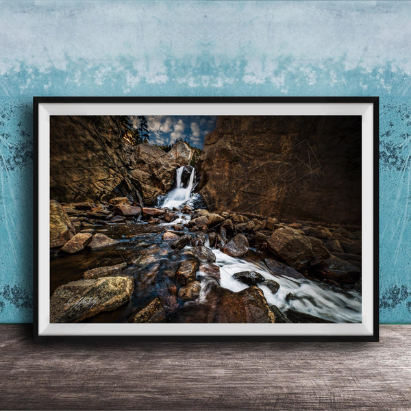 A framed photograph of a rocky waterfall cascading over rocks, set against a blue wall with a textured pattern.