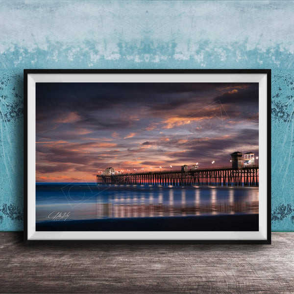 A framed photograph of a wooden pier extending into the ocean at sunset, with the sky filled with vibrant colors and clouds.