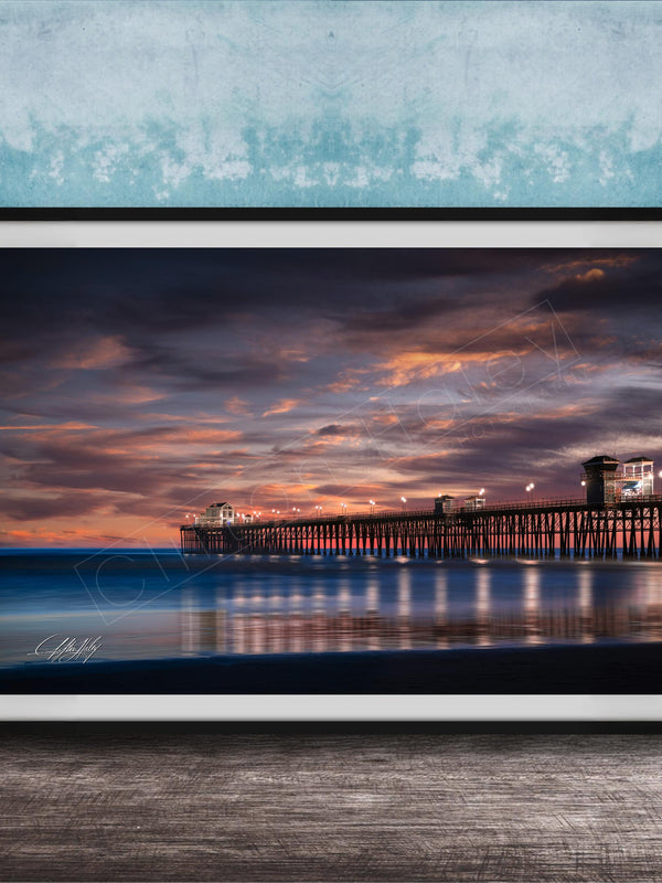 A framed photograph of a wooden pier extending into the ocean at sunset, with the sky filled with vibrant colors and clouds.