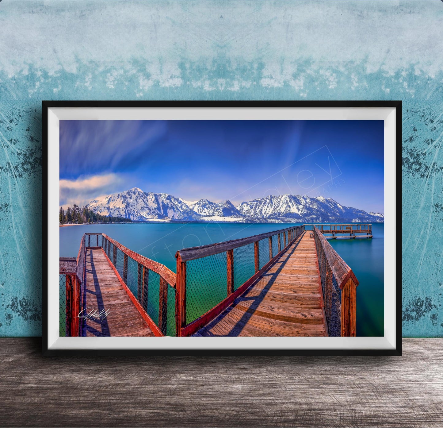 A framed photograph of a wooden pier extending into a serene lake, with snow-capped mountains in the background.