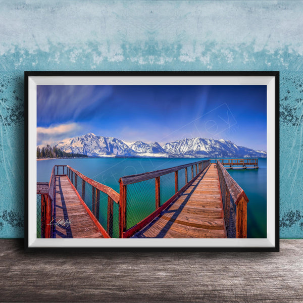 A framed photograph of a wooden pier extending into a serene lake, with snow-capped mountains in the background.