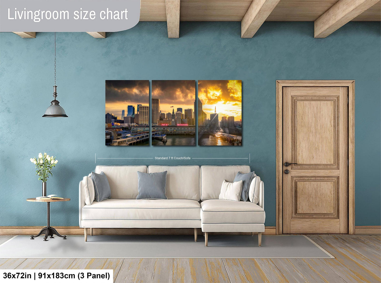 The image is a living room with a large wall art depicting a city skyline at sunset. The wall art is divided into three panels, each showing a different part of the cityscape. The room has a white sofa, a wooden door, and a wooden table with a vase of flowers. The wall art is approximately 36 inches wide and 9 inches tall, and it is mounted on a blue wall.