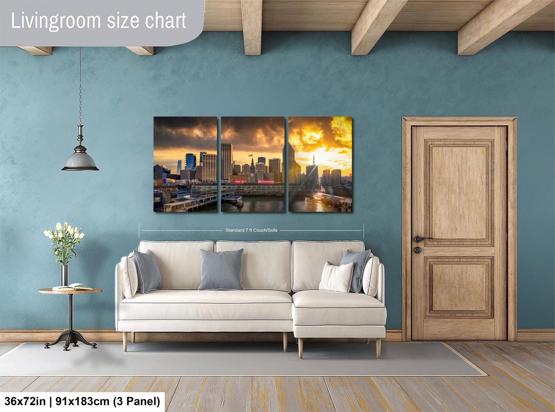 The image is a living room with a large wall art depicting a city skyline at sunset. The wall art is divided into three panels, each showing a different part of the cityscape. The room has a white sofa, a wooden door, and a wooden table with a vase of flowers. The wall art is approximately 36 inches wide and 9 inches tall, and it is mounted on a blue wall.