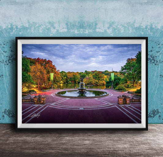 A framed photograph of a park with a fountain, surrounded by trees with autumn foliage, set against a blue wall.