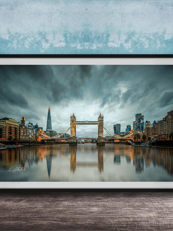 A framed photograph of the Tower Bridge in London, England, with the city's skyline reflected in the water below. The sky is filled with clouds, creating a dramatic atmosphere.