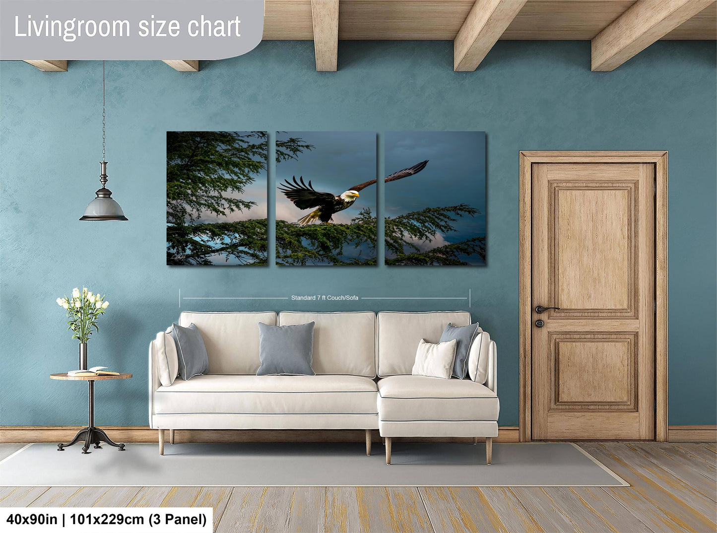 The image is a living room with a large white sofa, a wooden door, and a hanging light fixture. Above the sofa, there is a large wall art collage depicting a bald eagle in flight. The collage is composed of three panels, each showing the eagle soaring through the sky. The wall art is approximately 40 inches wide and 101.29 cm in height, with a 3-panel arrangement.