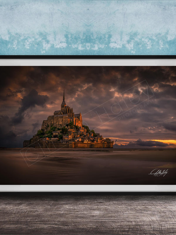 A framed photograph of the iconic Mont Saint-Michel in Normandy, France, with a dramatic sky in the background.