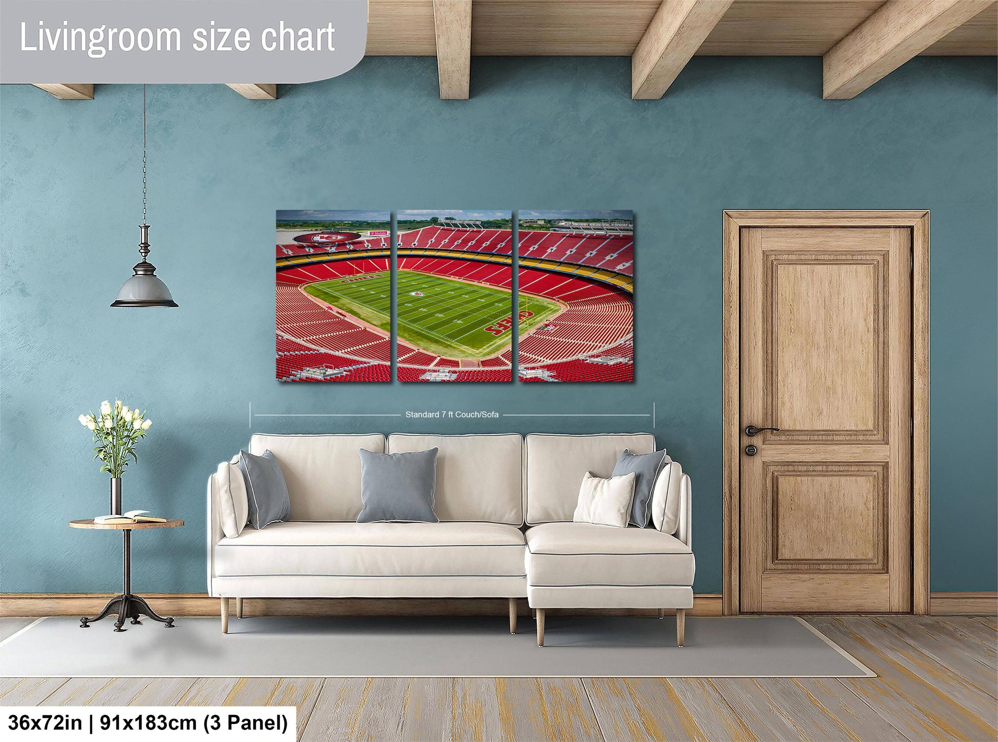 The image is a living room with a large wall-mounted painting depicting a stadium. The painting is divided into three panels, with the largest panel covering the entire wall. The room has a white sofa, a wooden door, and a small table with a vase of flowers. The painting is approximately 36 inches wide and 15 inches tall, and it is mounted on the wall above the sofa.