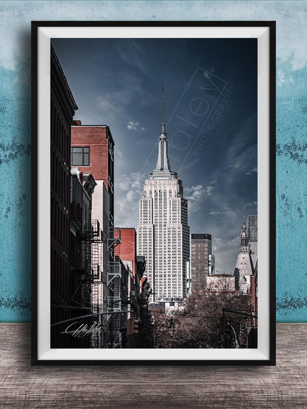 A framed photograph of the iconic Empire State Building in New York City, with a blue sky and clouds in the background.