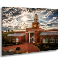 A large brick building with a prominent tower, surrounded by landscaping and set against a dramatic cloudy sky.