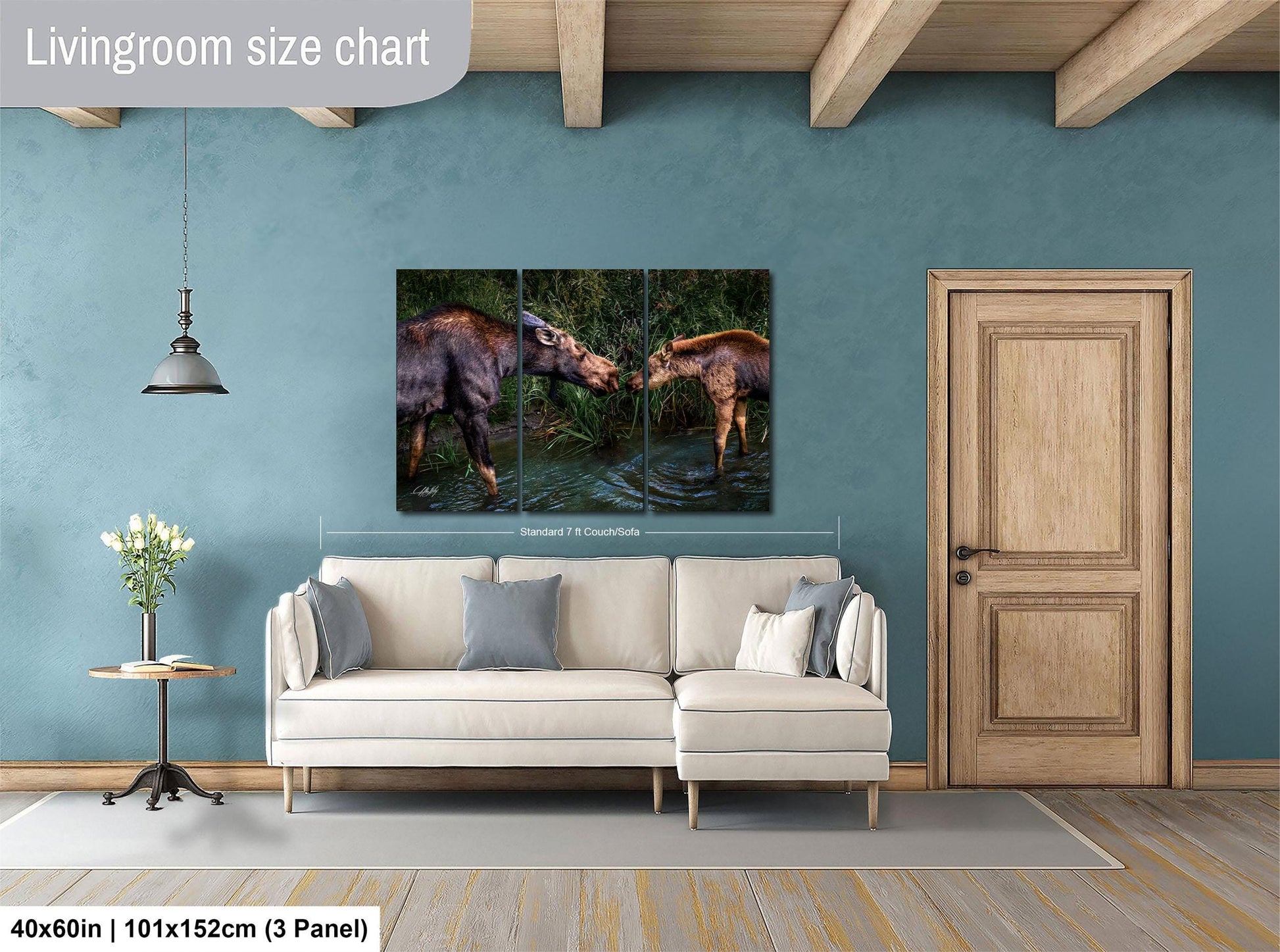 The image is a living room with a large painting of two moose in a pond hanging on the wall. The painting is framed and measures 40 inches by 101.5 inches, with a 3-panel layout. The room has a white sofa, a wooden door, and a small table with a vase of flowers.