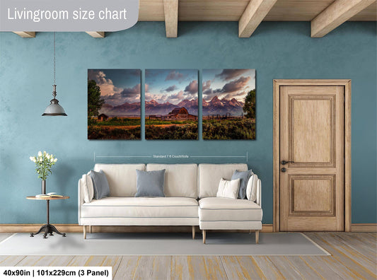 The image is a living room with a large, three-panel landscape painting hanging on the wall. The painting depicts a scenic mountain landscape with a sunset. The room has a white sofa and a wooden door, and there is a vase of flowers on a table. The painting is approximately 40 inches wide and 101.29 cm tall, with each panel measuring approximately 3 feet by 3.5 feet.