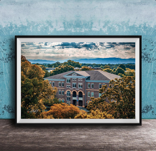 A framed photograph of a large brick building surrounded by trees with autumn foliage, set against a backdrop of mountains and a cloudy sky.