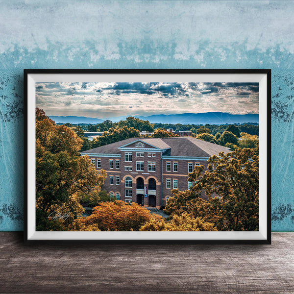 A framed photograph of a large brick building surrounded by trees with autumn foliage, set against a backdrop of mountains and a cloudy sky.