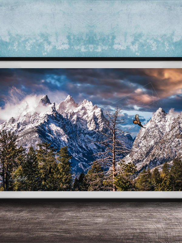 A framed photograph of a snow-capped mountain range with a cloudy sky, surrounded by pine trees and a lone bird in flight.
