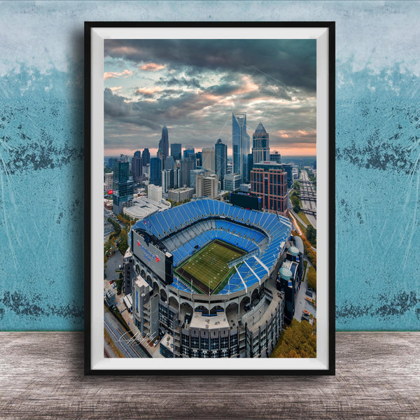An aerial view of a large sports stadium with a city skyline in the background, framed in a black frame.