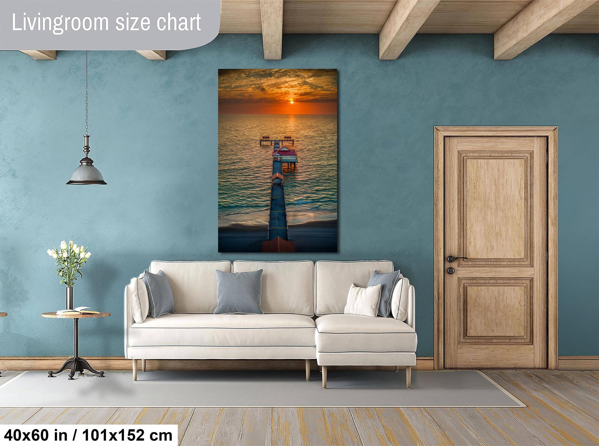 a living room with a large wall painting of a sunset over the ocean, a white sofa, a wooden door, and a small table with a vase of flowers.