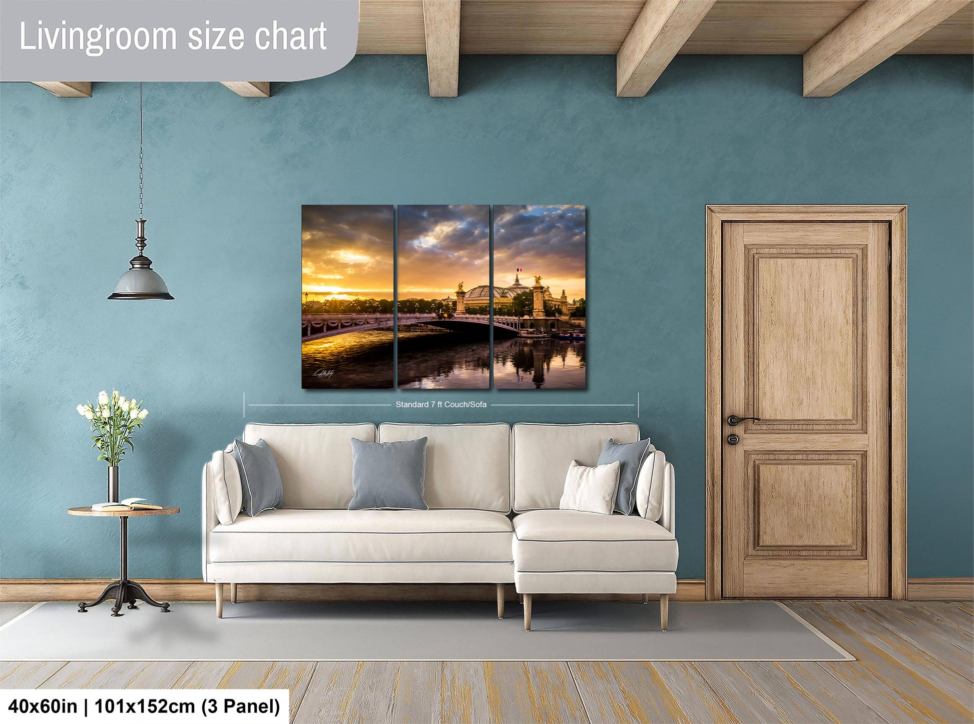 The image is a living room with a large, three-panel wall art depicting a scenic landscape with a bridge and a sunset. The wall art is mounted above a white sofa, and there is a wooden door and a small table with a vase of flowers nearby. The living room has a blue wall and a wooden floor, creating a cozy and inviting atmosphere.