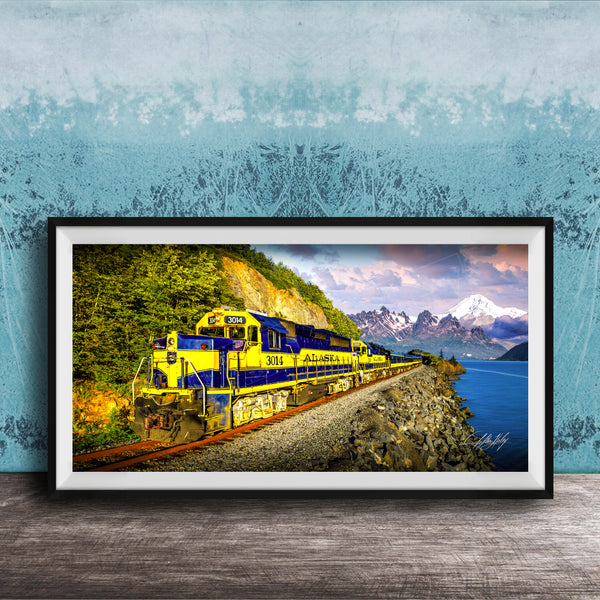 A framed photograph of a yellow and blue train traveling along a track through a mountainous landscape with a body of water in the background.