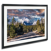 A framed photograph depicting a winding road surrounded by a forested mountain landscape with snow-capped peaks in the distance.