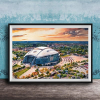 The image is a framed painting depicting a large stadium with a curved roof, set against a backdrop of a city skyline and a colorful sunset sky.