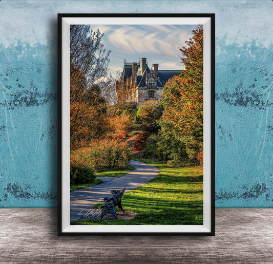 A framed photograph of a park with a winding path, a bench, and trees with autumn foliage. In the background, there is a large, ornate building with a gabled roof and multiple chimneys.