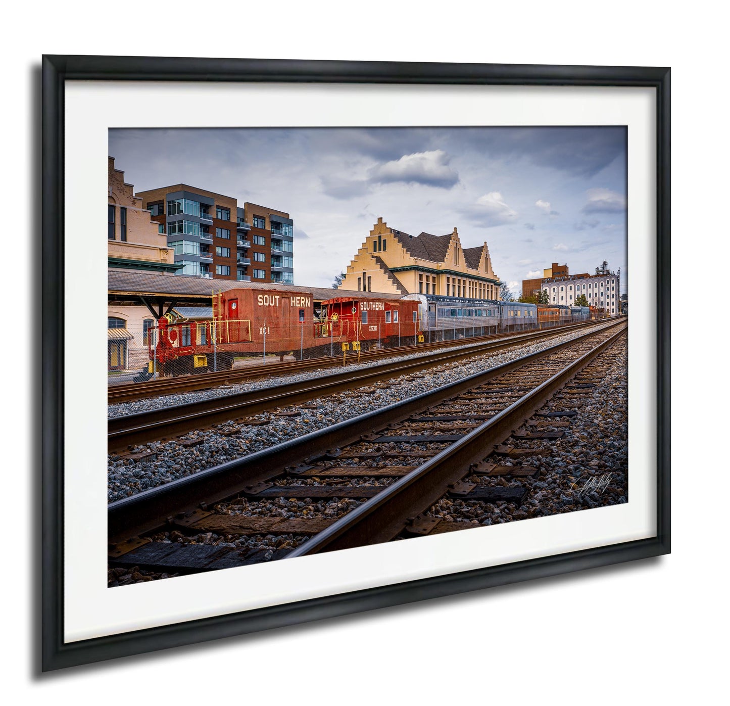 A framed photograph depicting a train traveling along train tracks, with buildings and a cloudy sky in the background.
