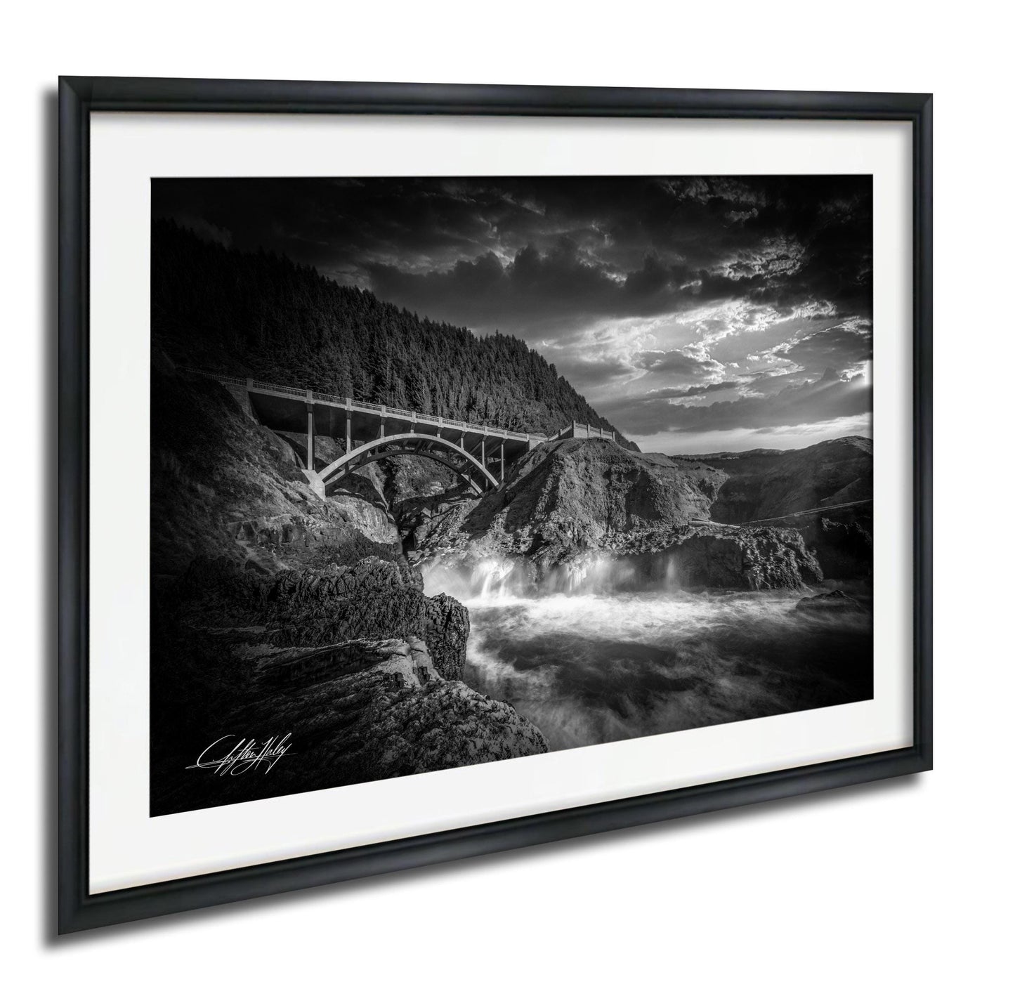 A black and white photograph of a bridge spanning over a river, with a dramatic sky and waterfalls in the background.