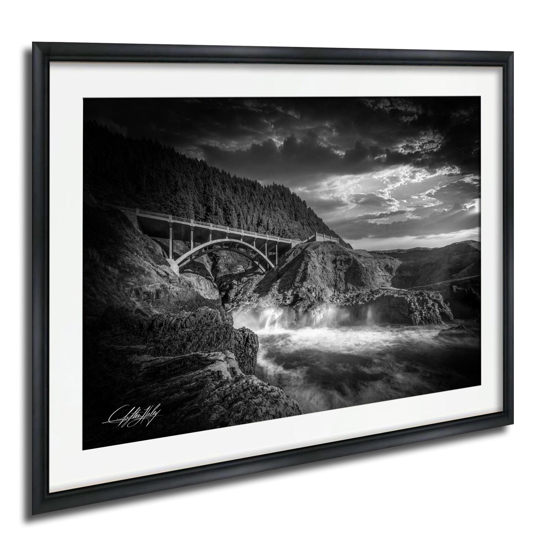 A black and white photograph of a bridge spanning over a river, with a dramatic sky and waterfalls in the background.
