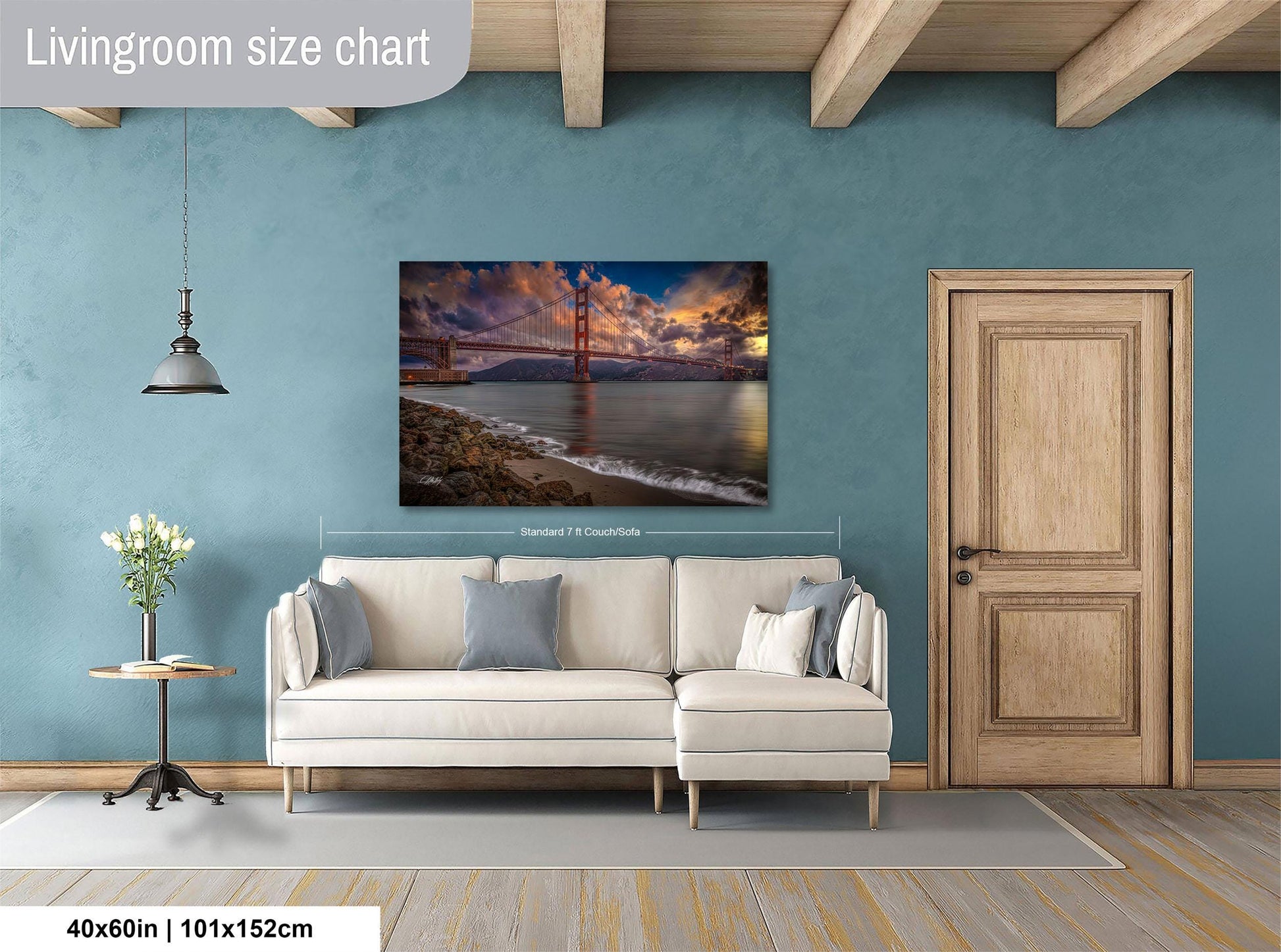 a living room with a large wall painting depicting a scenic view of the Golden Gate Bridge at sunset. The painting is framed and hangs above a white sofa, which is positioned in front of a wooden door. The room has a wooden floor and a small table with a vase of flowers. The wall painting is approximately 40 inches wide and 115 centimeters tall.